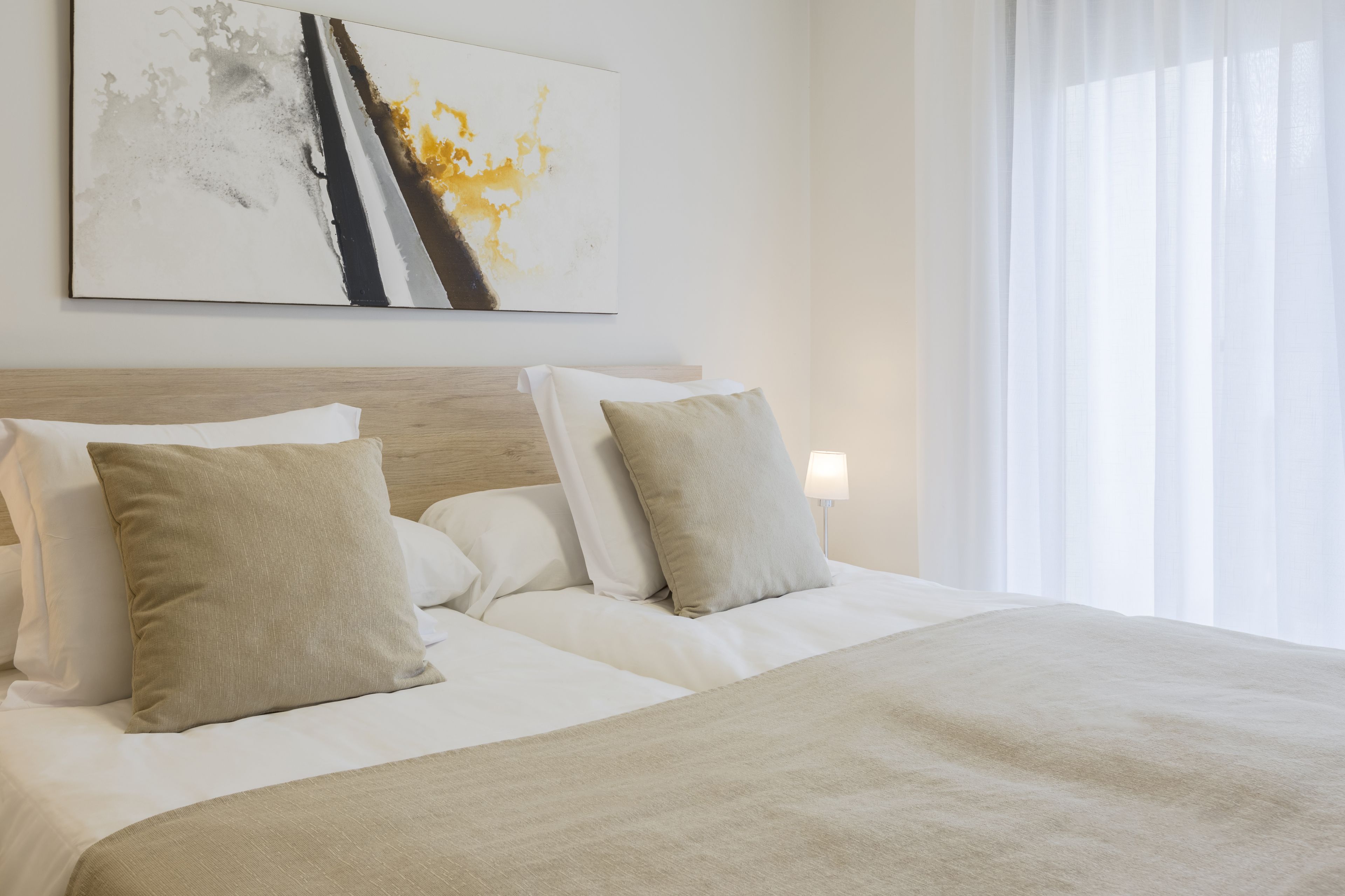 Ein ordentlich bezogenes Bett mit beige-farbener Bettwäsche und Kissen, ein hölzernes Kopfteil und ein großes abstraktes Gemälde an der Wand.