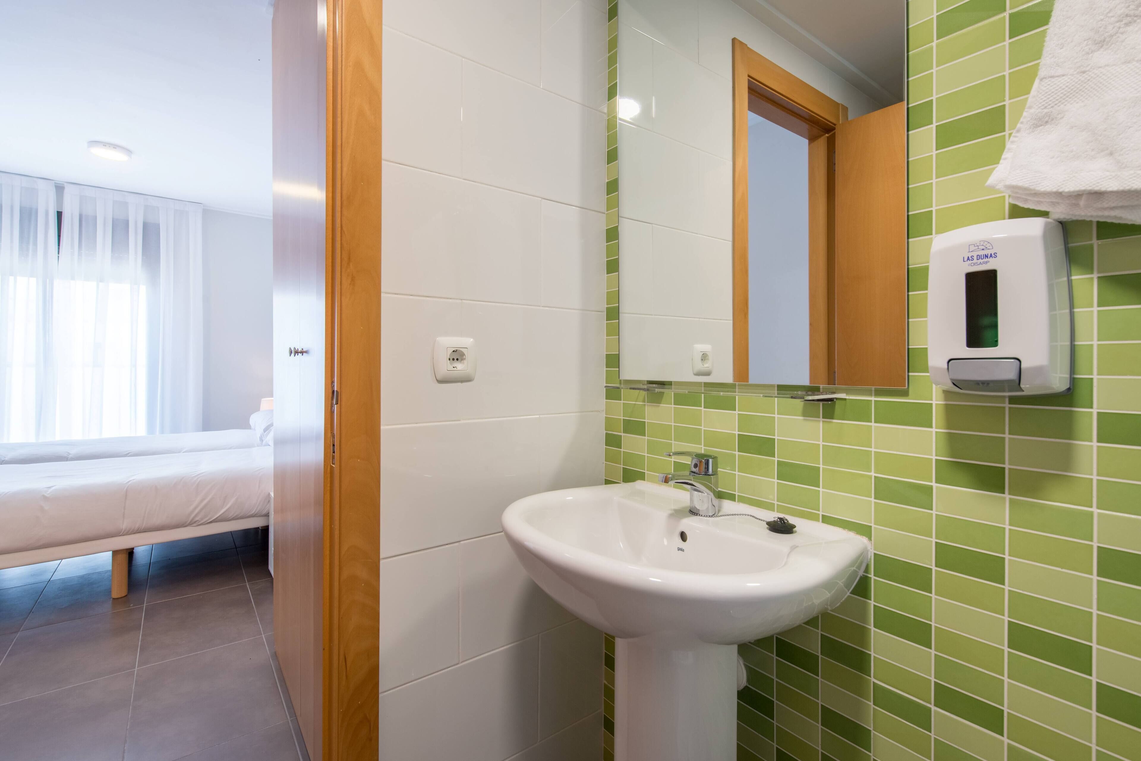 Un baño con un lavabo blanco, paredes cubiertas de azulejos verdes, un espejo y un dispensador de gel antibacterial.