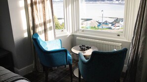 Ein Zimmer mit zwei blauen Stühlen, einem kleinen Tisch und einem Fenster mit Blick auf ein Gewässer und Gebäude.