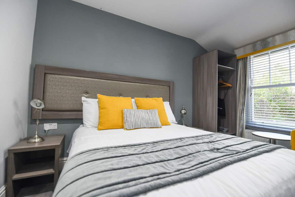 Un lit bien fait, recouvert d’une couverture à rayures grises et blanches, avec deux oreillers jaunes et une tête de lit grise.