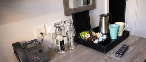 A hotel room with a desk featuring a Voitelink phone, a coffee maker, two cups, a remote control, and a tray with snacks and a water bottle.