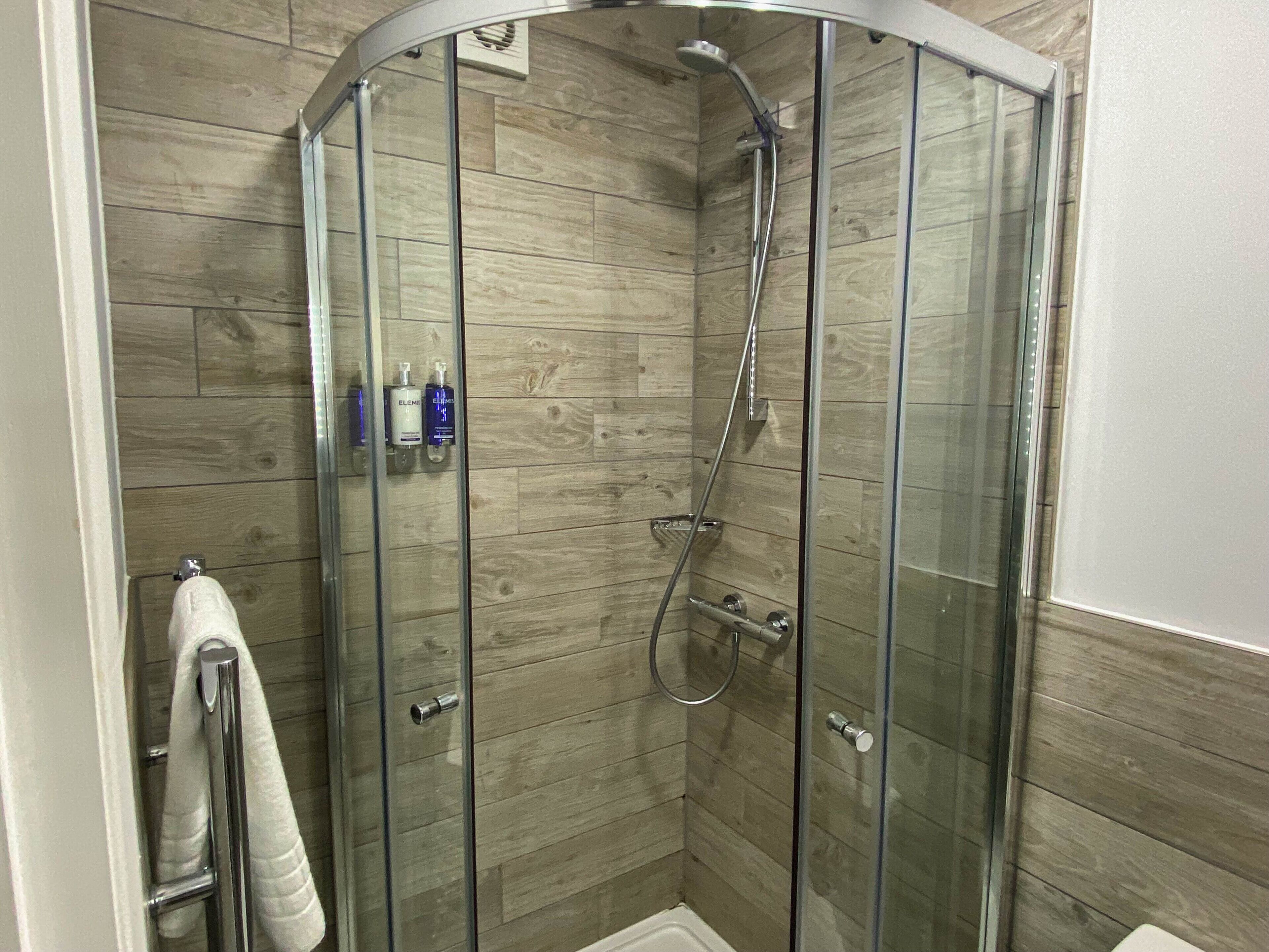 A glass-enclosed shower with a wooden panel wall, a towel rack, and two bottles of Elemis body wash.
