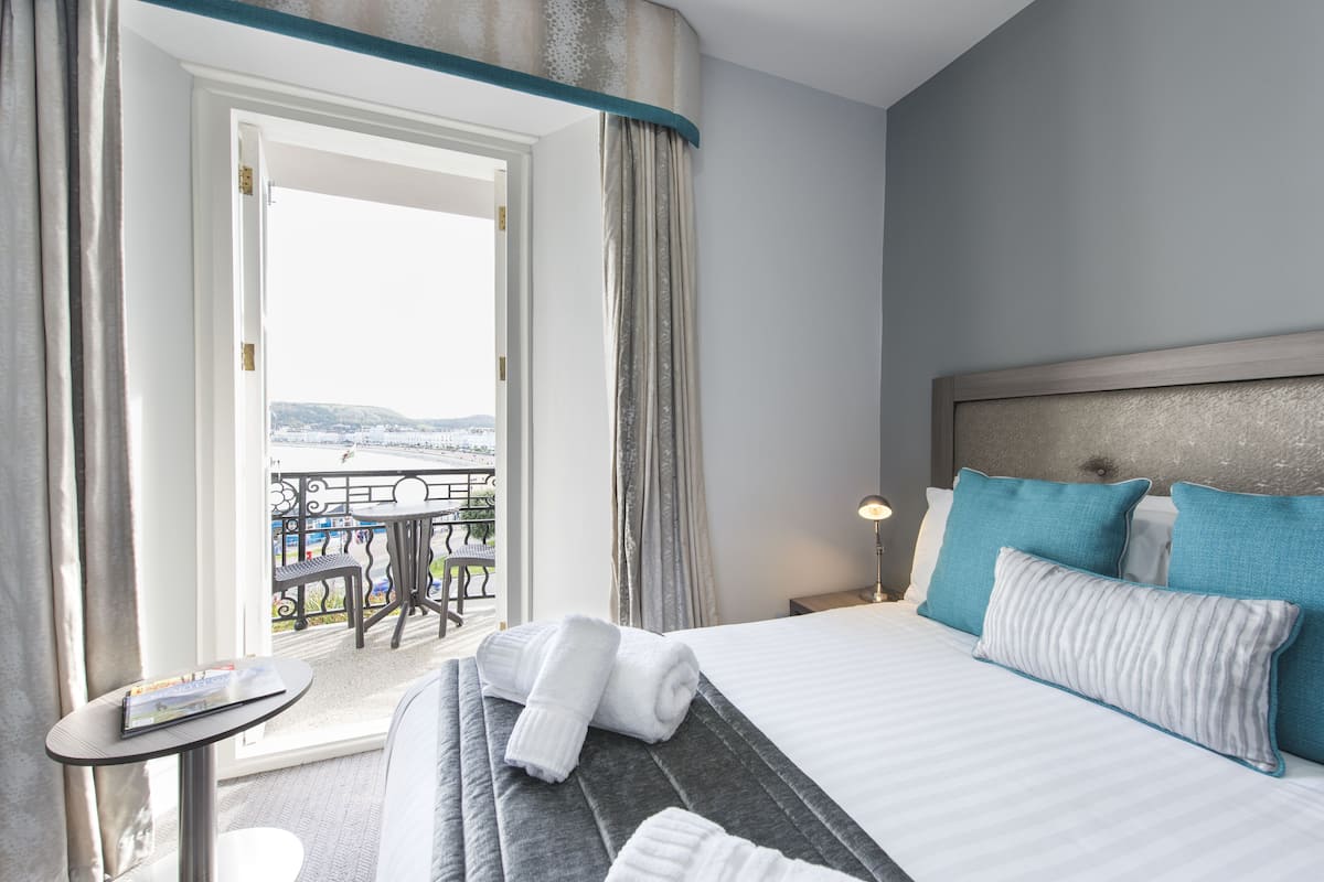 Une chambre d’hôtel avec un lit, une petite table avec un magazine, un balcon avec une table et des chaises, et une vue sur la mer.
