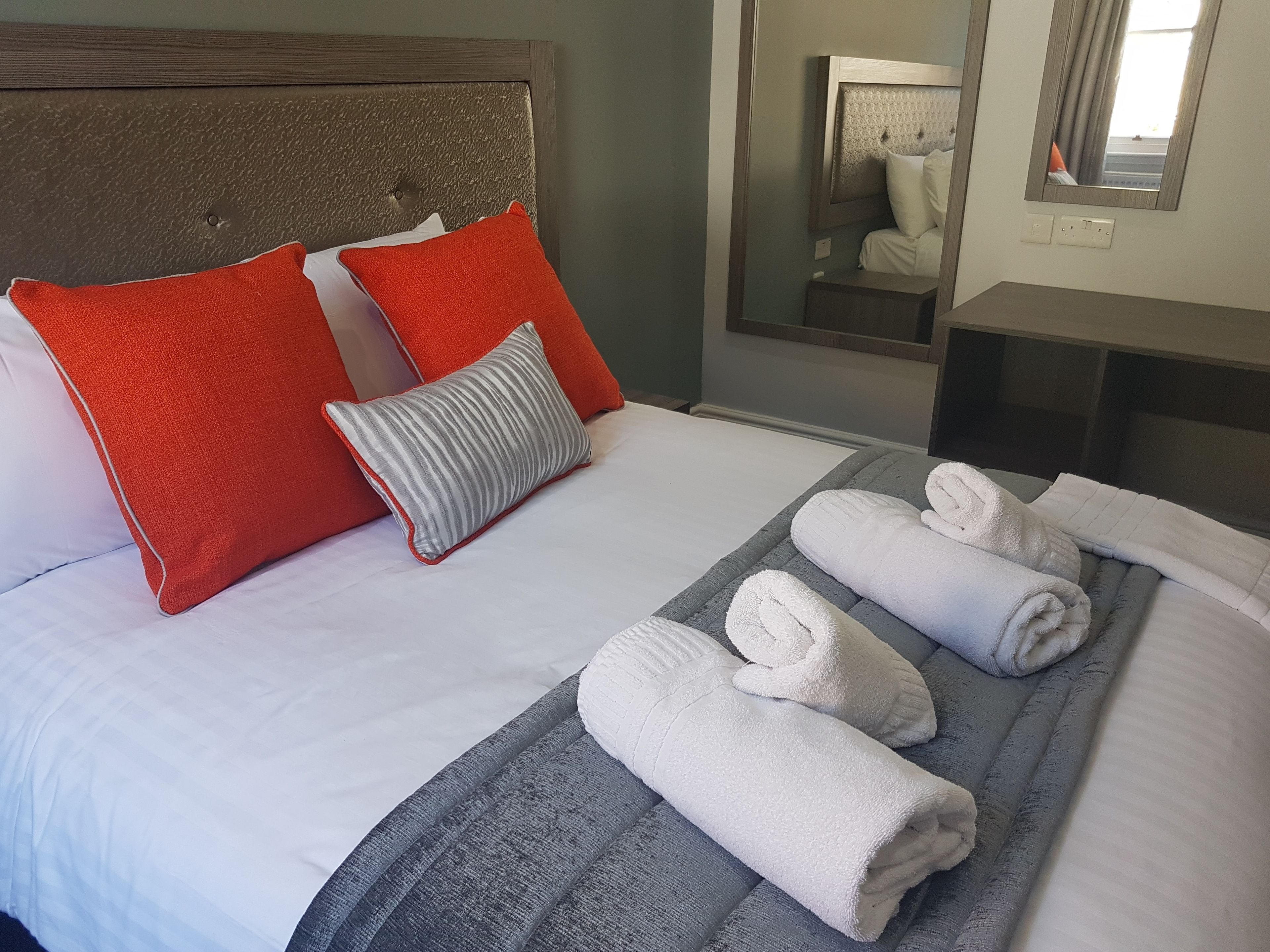 A neatly made bed with white and red pillows, a grey striped pillow, and rolled white towels on a grey blanket.
