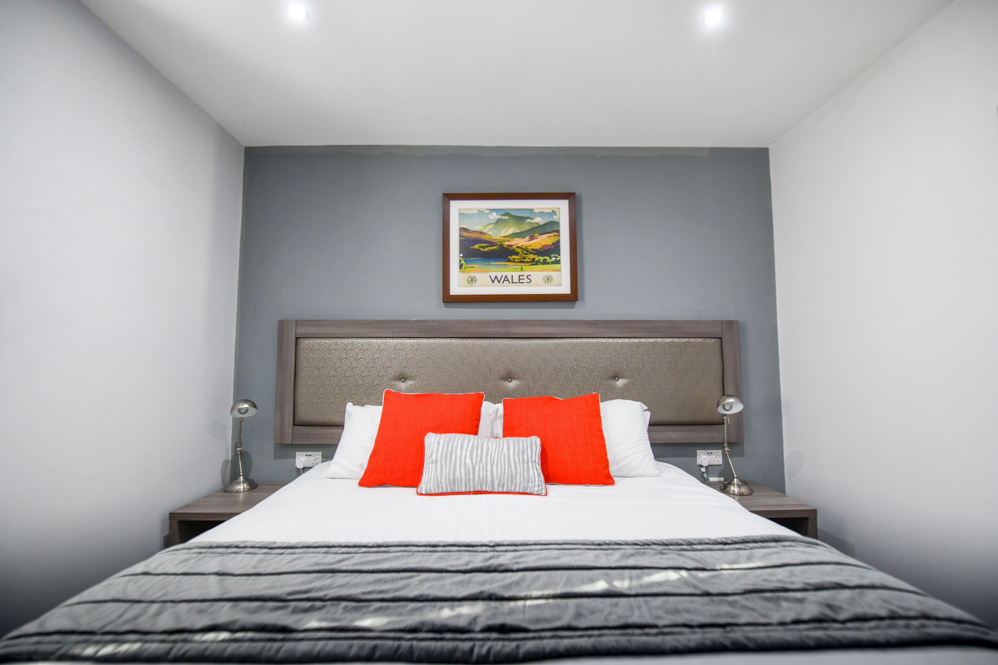 A neatly made bed with white and red pillows, a framed picture of Wales on the wall, and a bedside lamp on each side.
