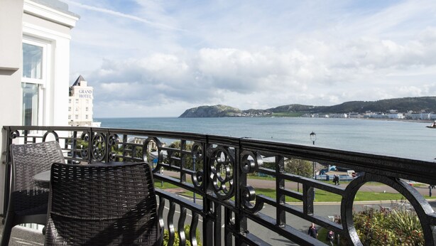 Ein Balkon mit Korbmöbeln, der auf ein Gewässer, ein entferntes Gebäude und einen klaren Himmel blickt.