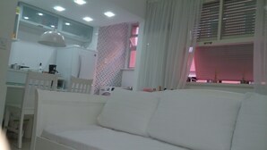 A white sofa in a room with a kitchen area, a dining table, and a window with blinds. - Estúdio Praia do Flamengo - 1018 (Rio de Janeiro)