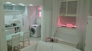 A compact, well-lit room with a kitchenette, a washing machine, a dining area, and a window with blinds. - Estúdio Praia do Flamengo - 1018 (Rio de Janeiro)