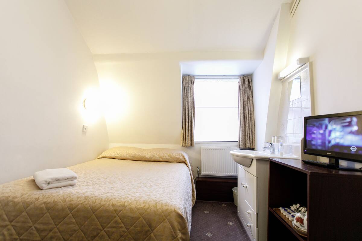 A hotel room with a bed, a small desk with a television, a window with curtains, and a radiator.
