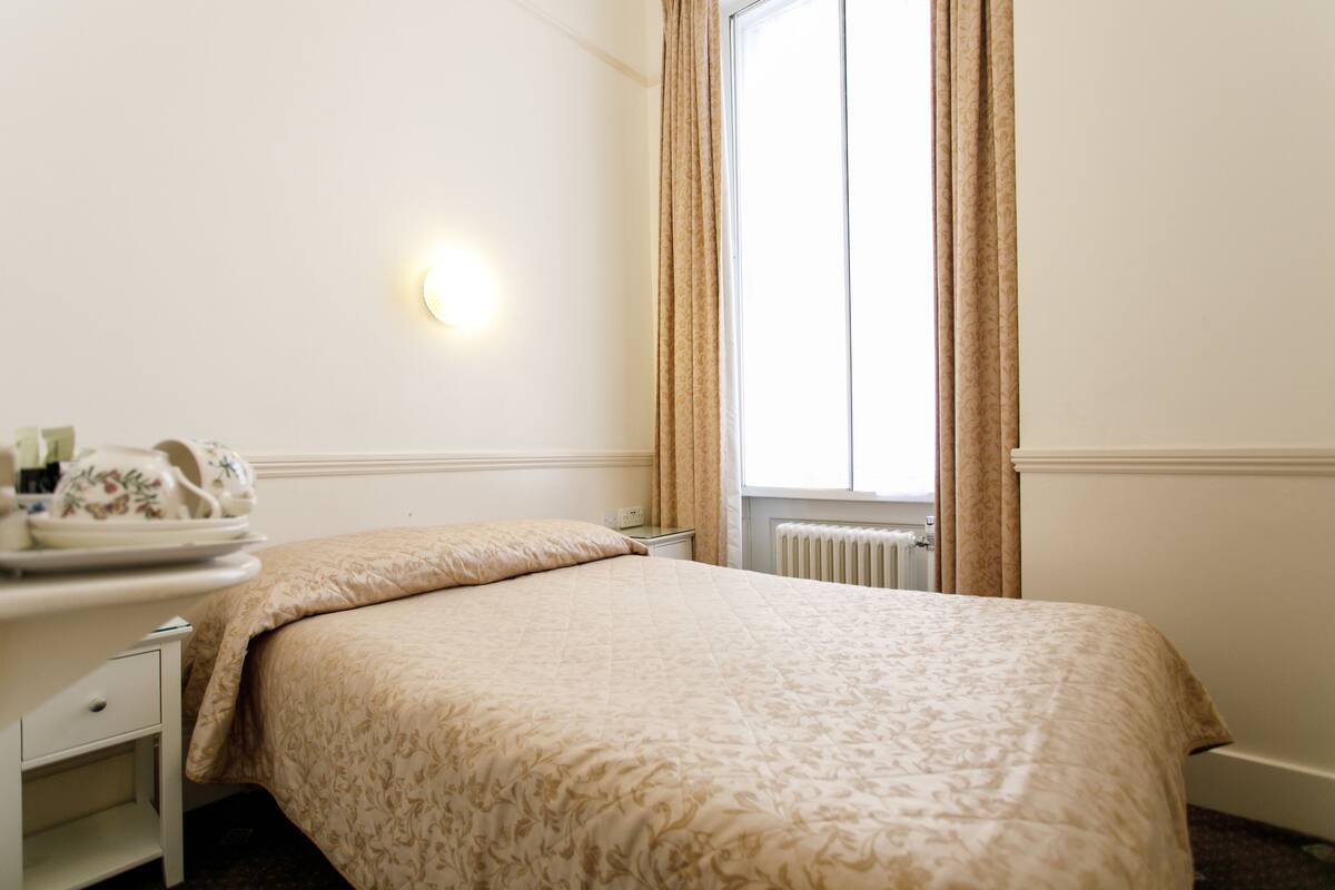 A hotel room with a bed, a bedside table with a lamp, and a window with curtains.