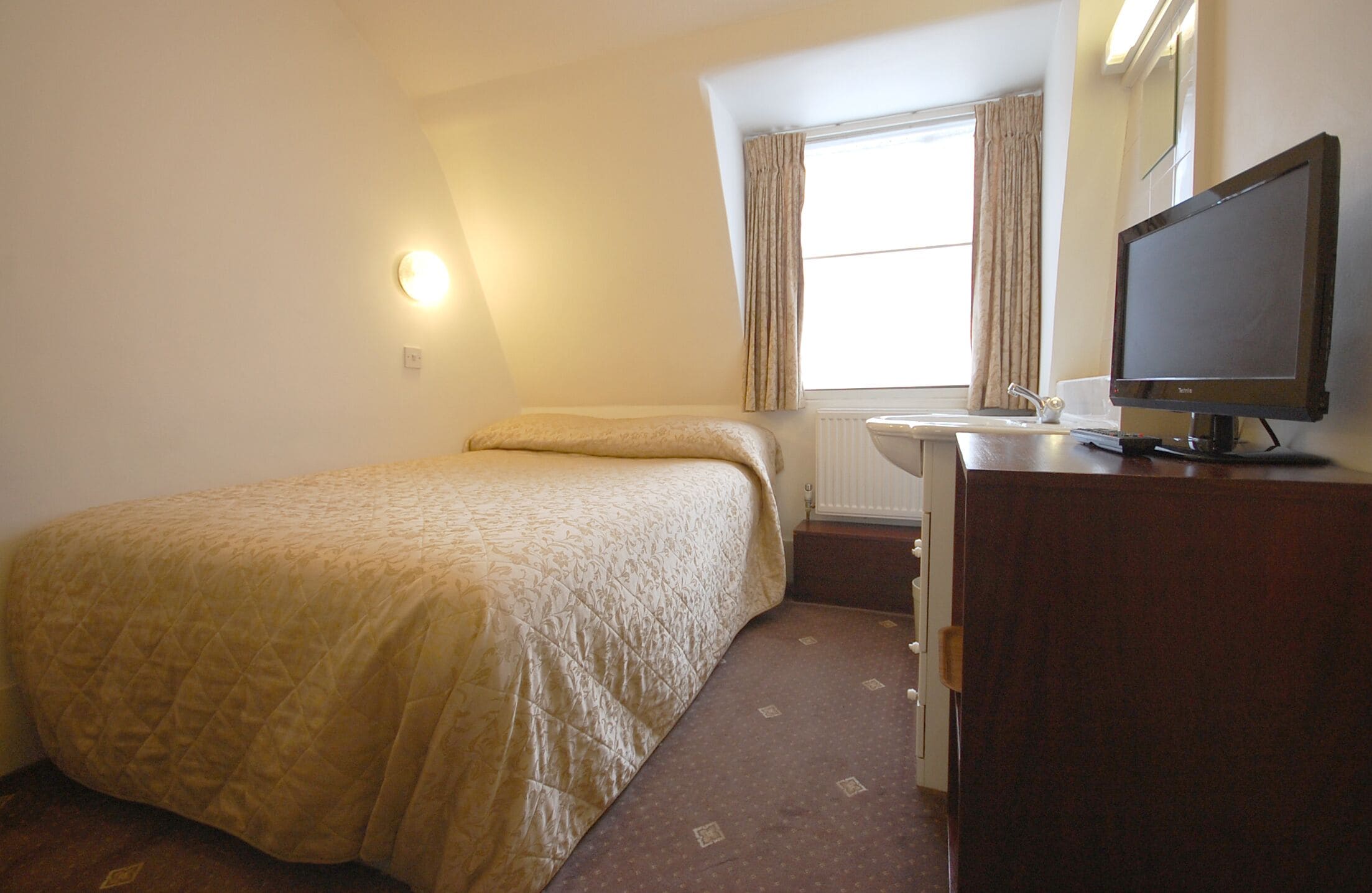 A small, neatly arranged bedroom with a bed, a dresser, a television, and a window with curtains.