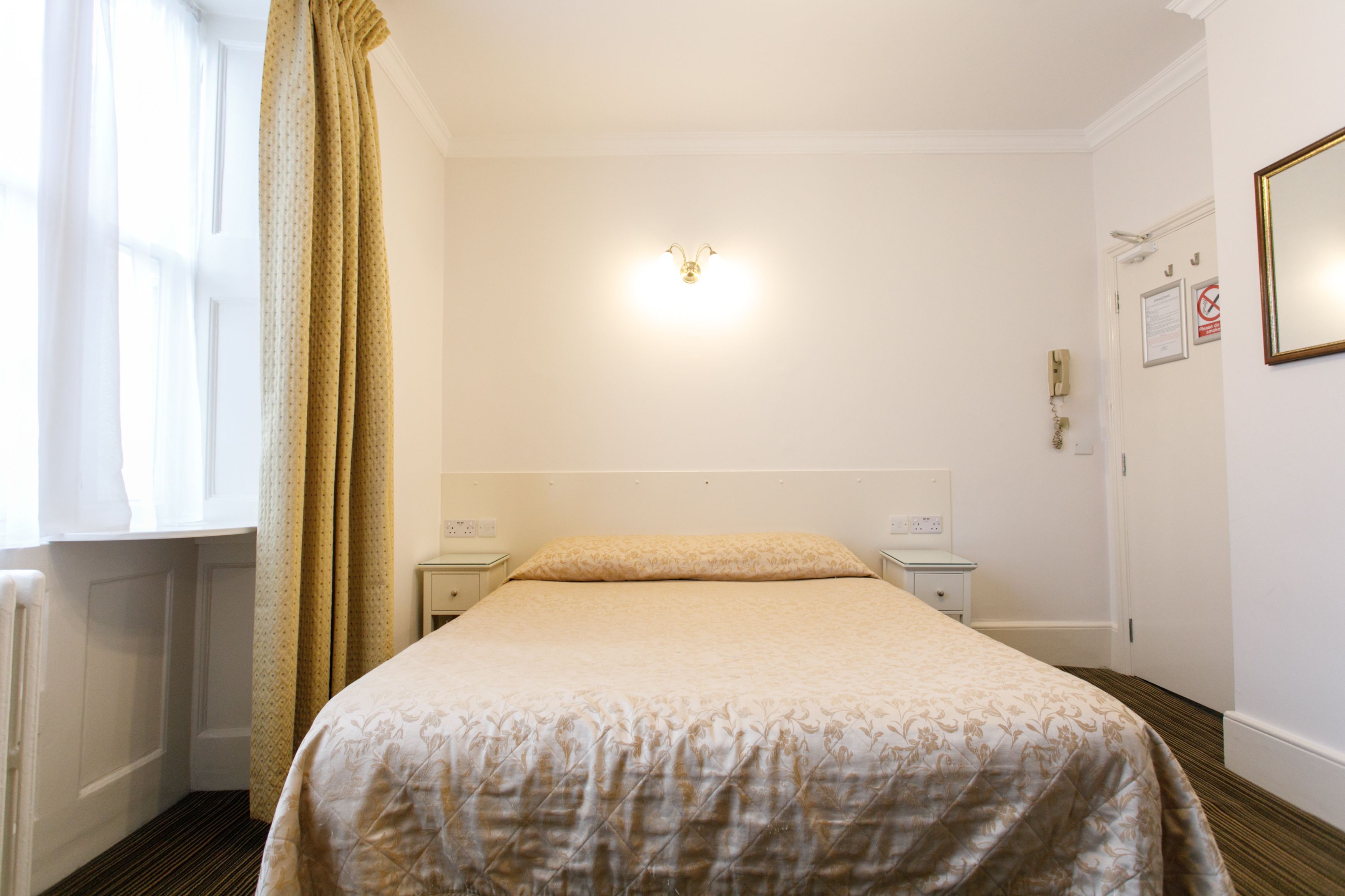 Una habitación de hotel con una cama, una mesita de noche, una lámpara de pared, una puerta con un cartel y una ventana con cortinas.