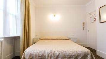 Una habitación de hotel con una cama, una mesita de noche, una lámpara de pared, una puerta con un cartel y una ventana con cortinas.