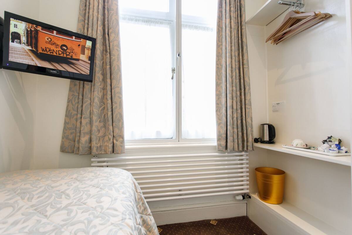 A bedroom with a bed, a window with curtains, a radiator, a shelf with items, and a wall-mounted TV displaying an image.