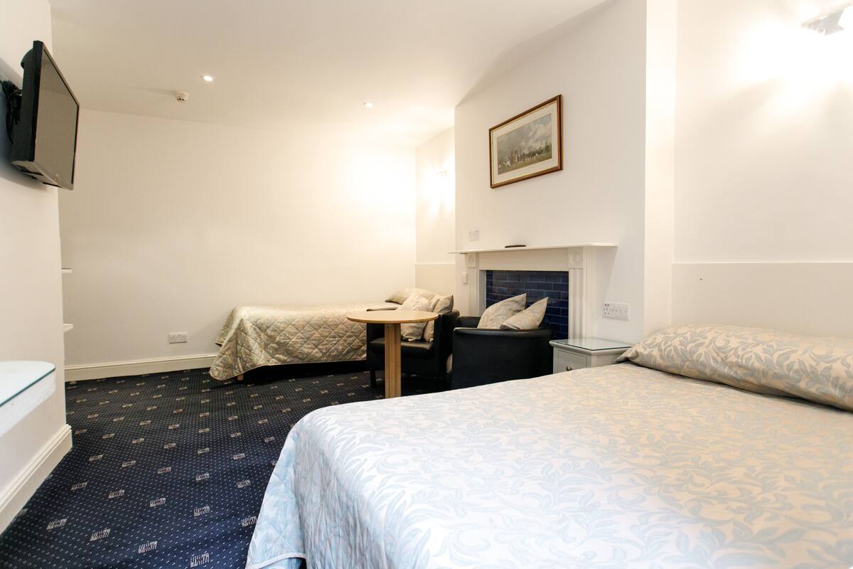 A hotel room with a bed, two single beds, a desk, a chair, a TV, and a fireplace.