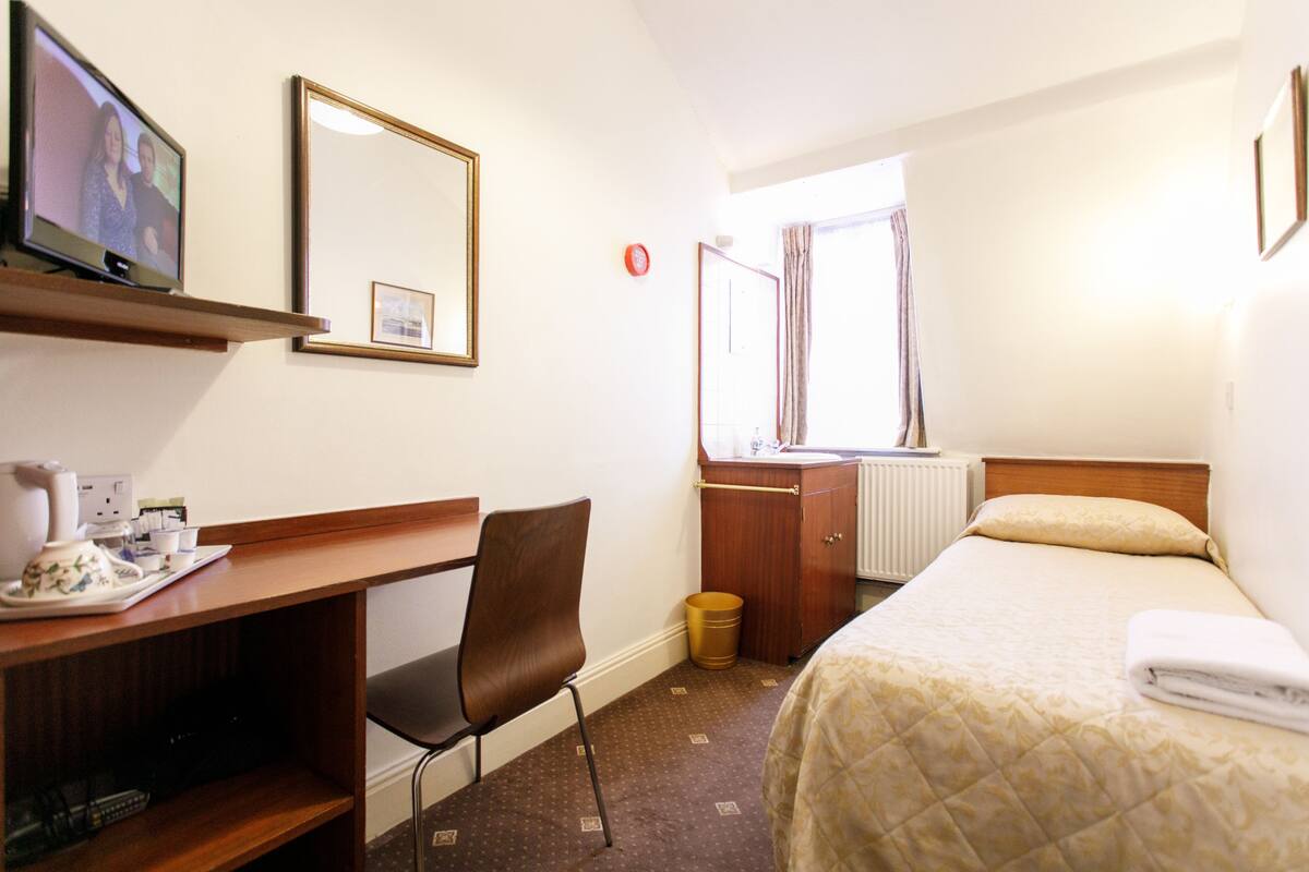 A hotel room with a bed, a desk with a chair, a television, a window with curtains, and a radiator.