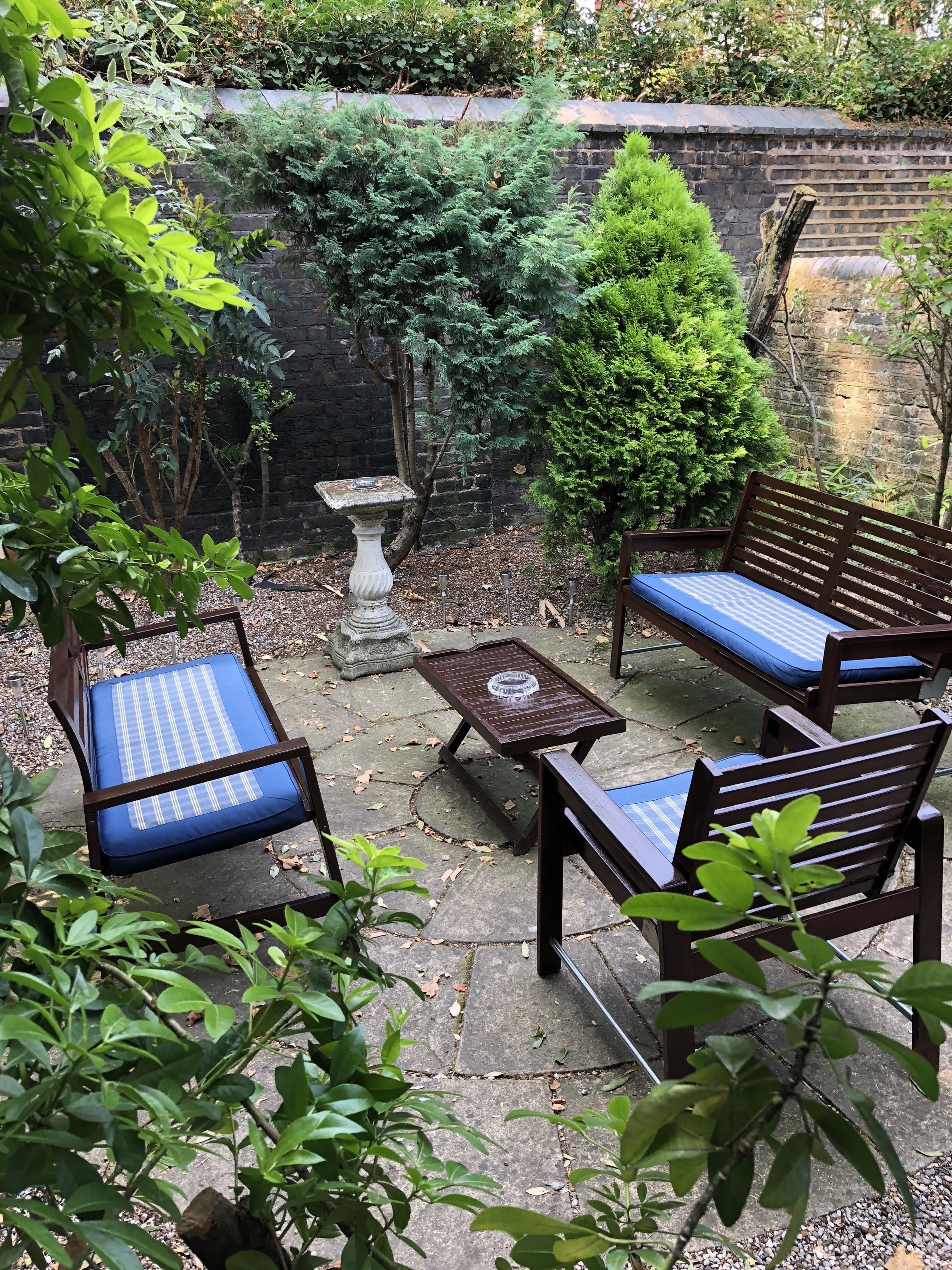 A patio area with two wooden chairs and a small table, surrounded by greenery and a brick wall.