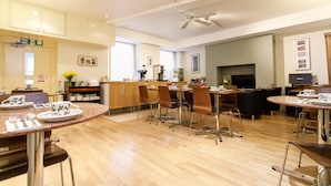 A spacious dining area with multiple tables and chairs, a kitchen area with a coffee machine, and a television in the background. - Jesmond Hotel (London)
