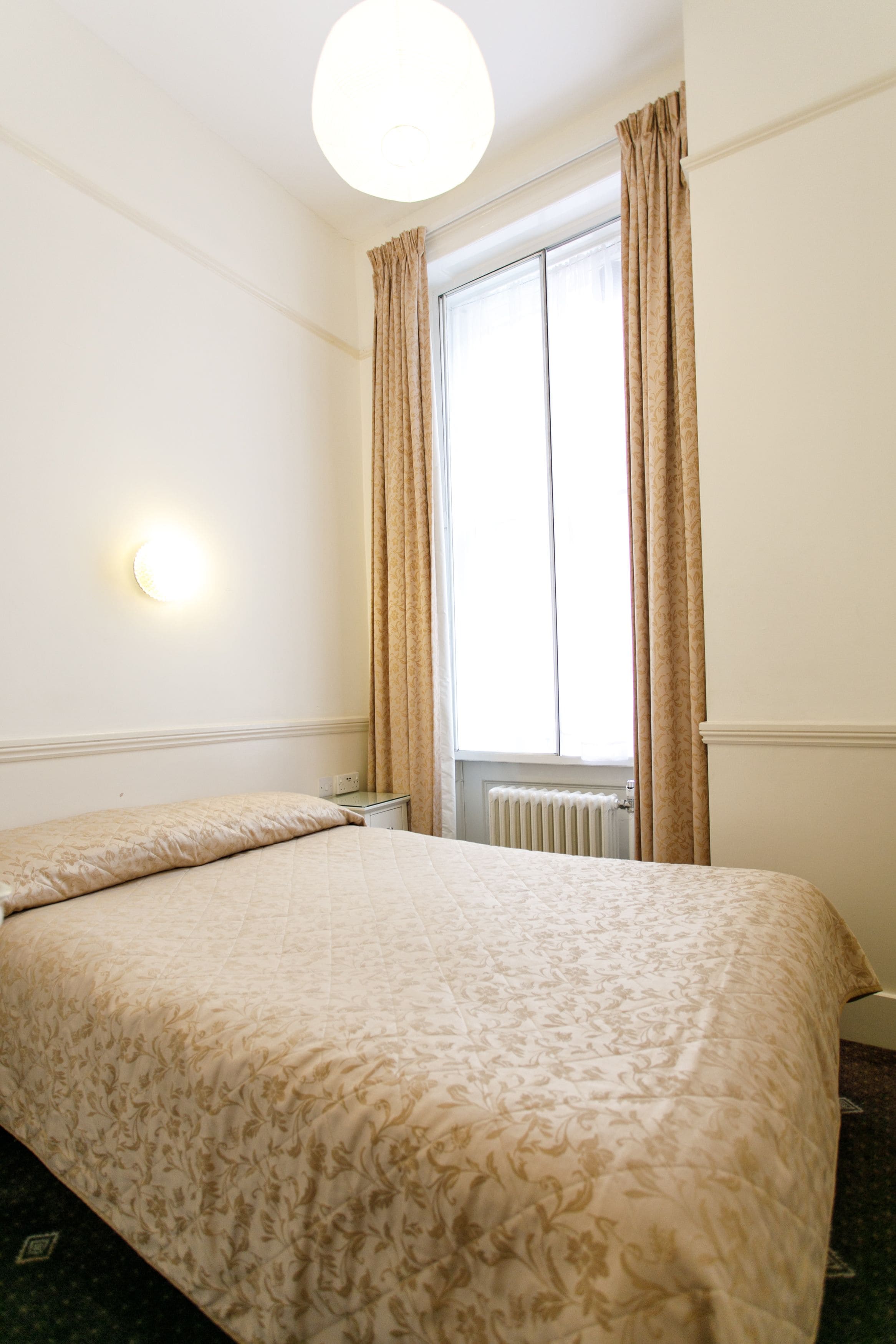 A hotel room with a bed, a nightstand, a wall-mounted light, and a window with curtains.