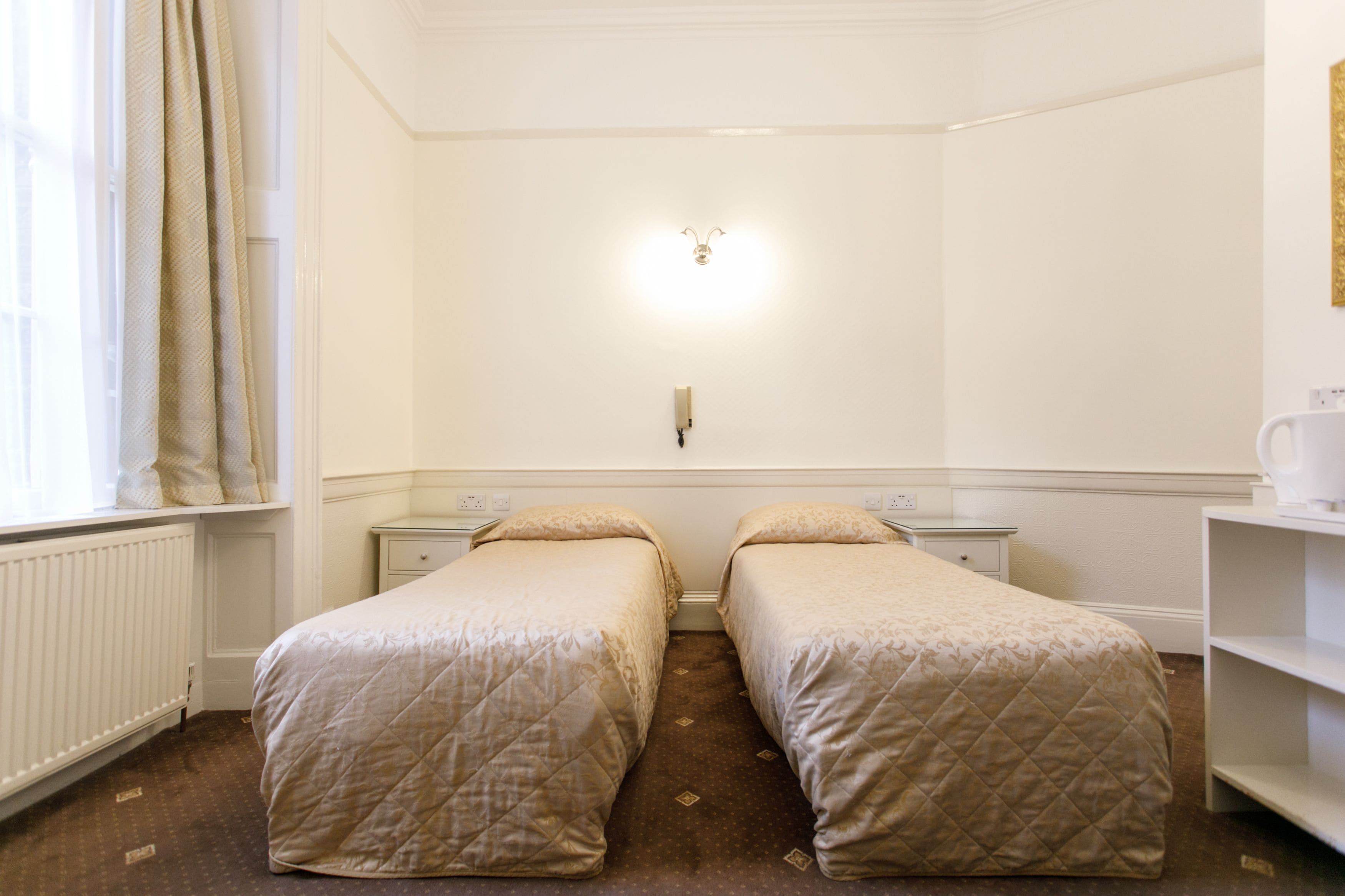 Habitación doble con dos camas individuales, una mesita de noche, un estante y una ventana con cortinas.
