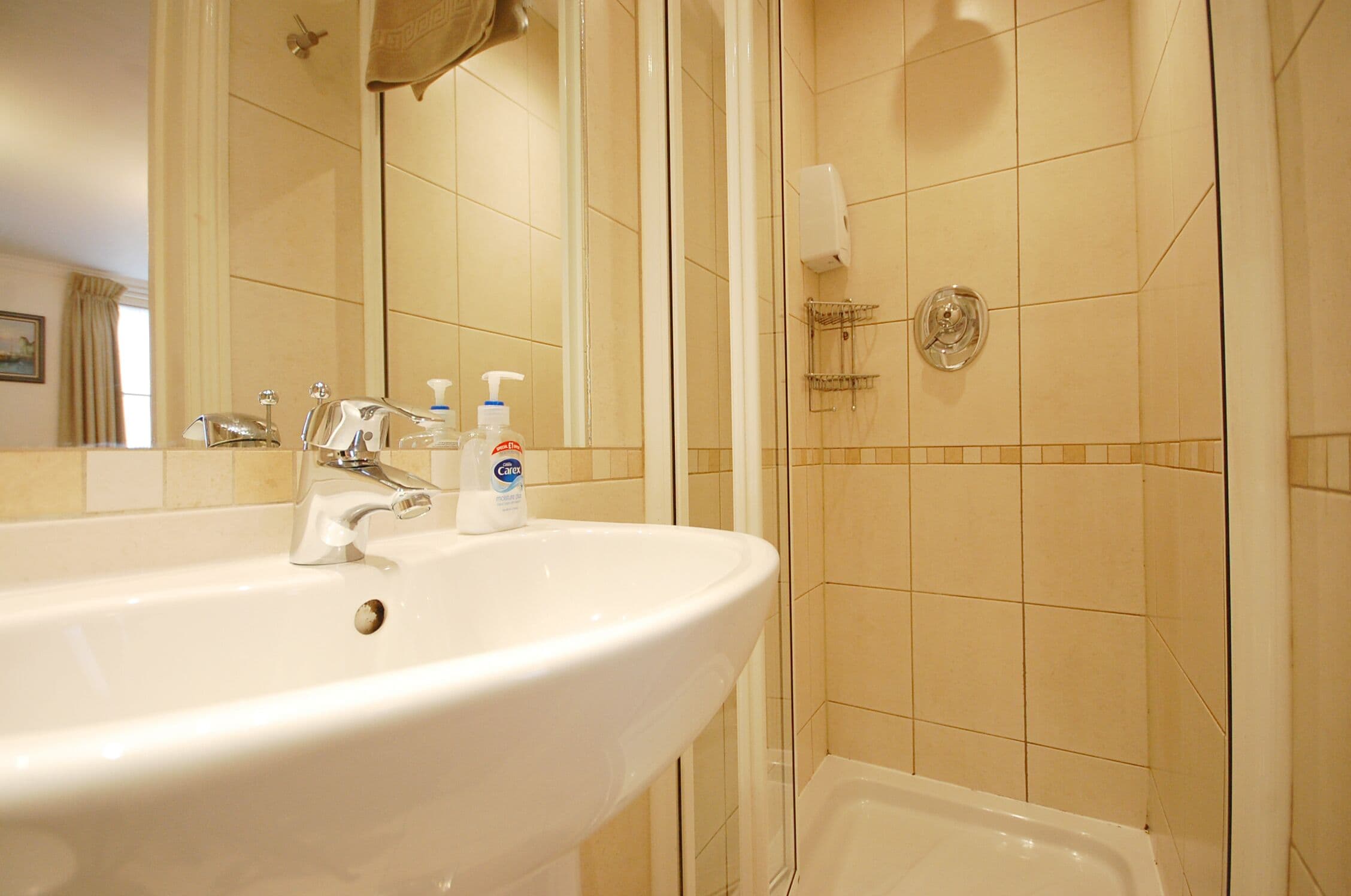 Un baño con lavamanos blanco, un espejo, dispensador de jabón y zona de ducha.