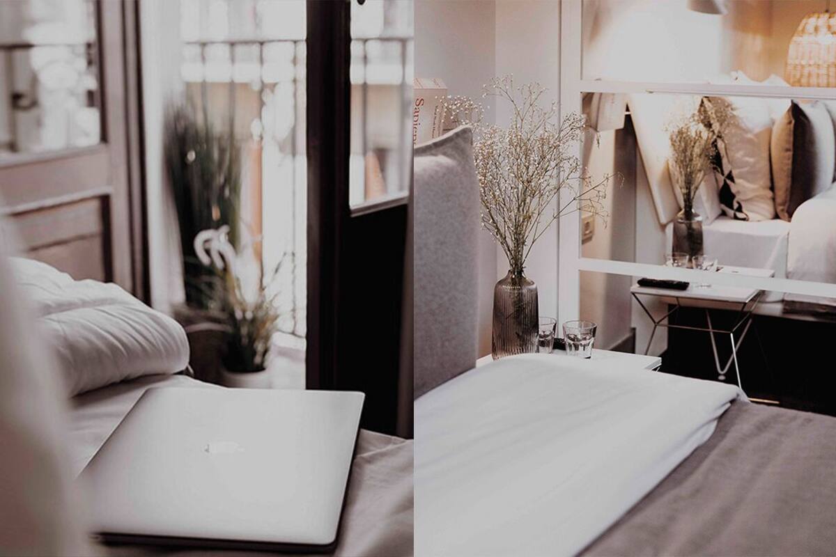 Egy rendezett hálószoba, az ágyon egy laptop, az éjjeliszekrényen egy száraz virágokból álló vázában, és az ablakon keresztül a külső tájra nyíló kilátás.