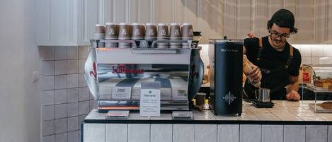 Interno di un caffè con lavagna dei prezzi, macchina per espresso e un barista che prepara delle bevande.