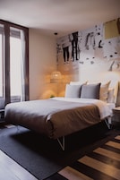 Ein modernes Schlafzimmer mit einem großen Bett, einem Nachttisch, einer Wandlampe und einem großen Fenster mit Vorhängen.