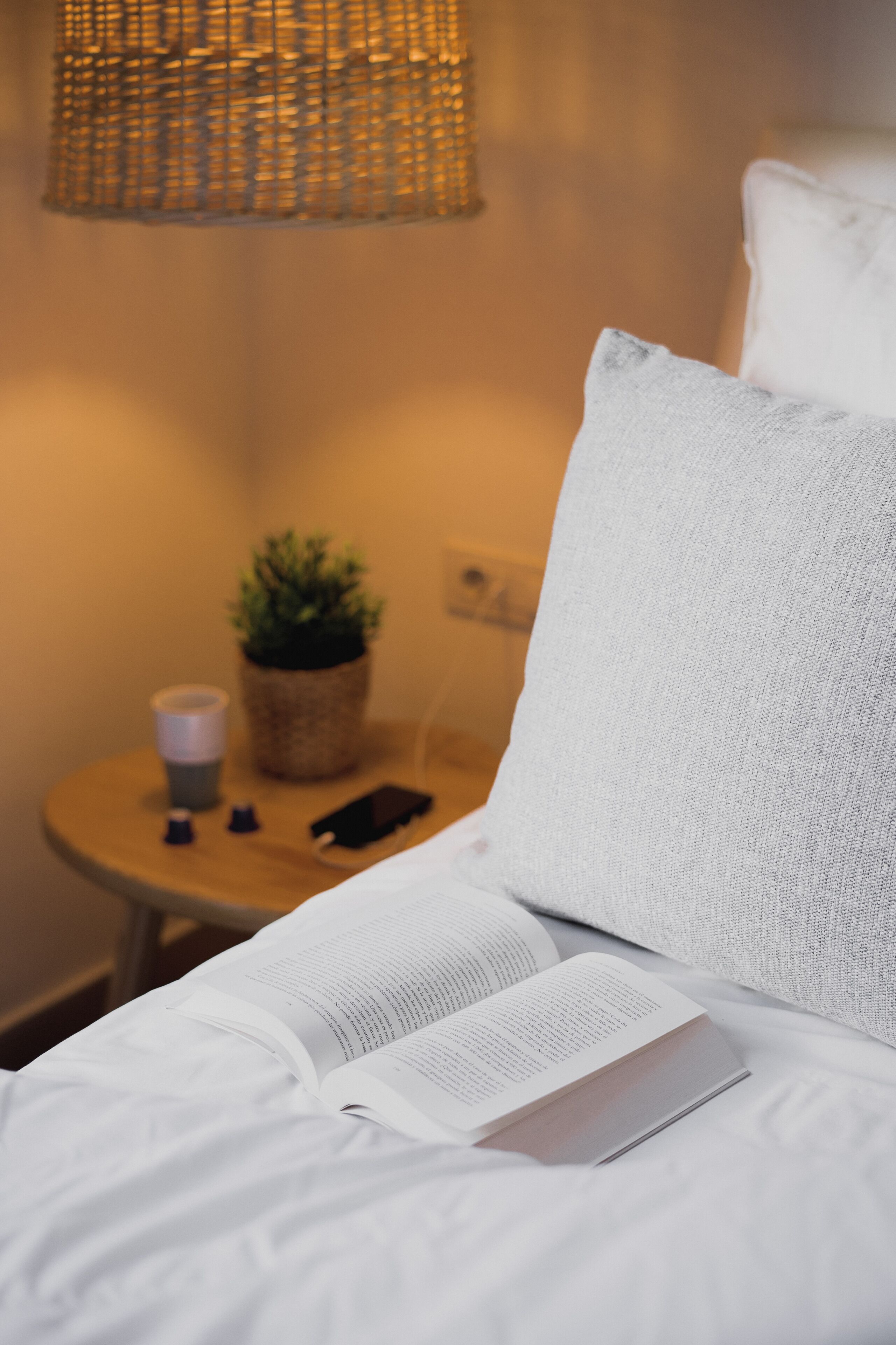 A neatly made bed with a book, a small potted plant, and a cup on a bedside table.