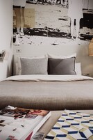 Ein ordentlich bezogenes Bett in Grau-Weiß gehalten, eine Zeitschrift daneben, und eine Wand mit einem großen, abstrakten Kunstwerk.