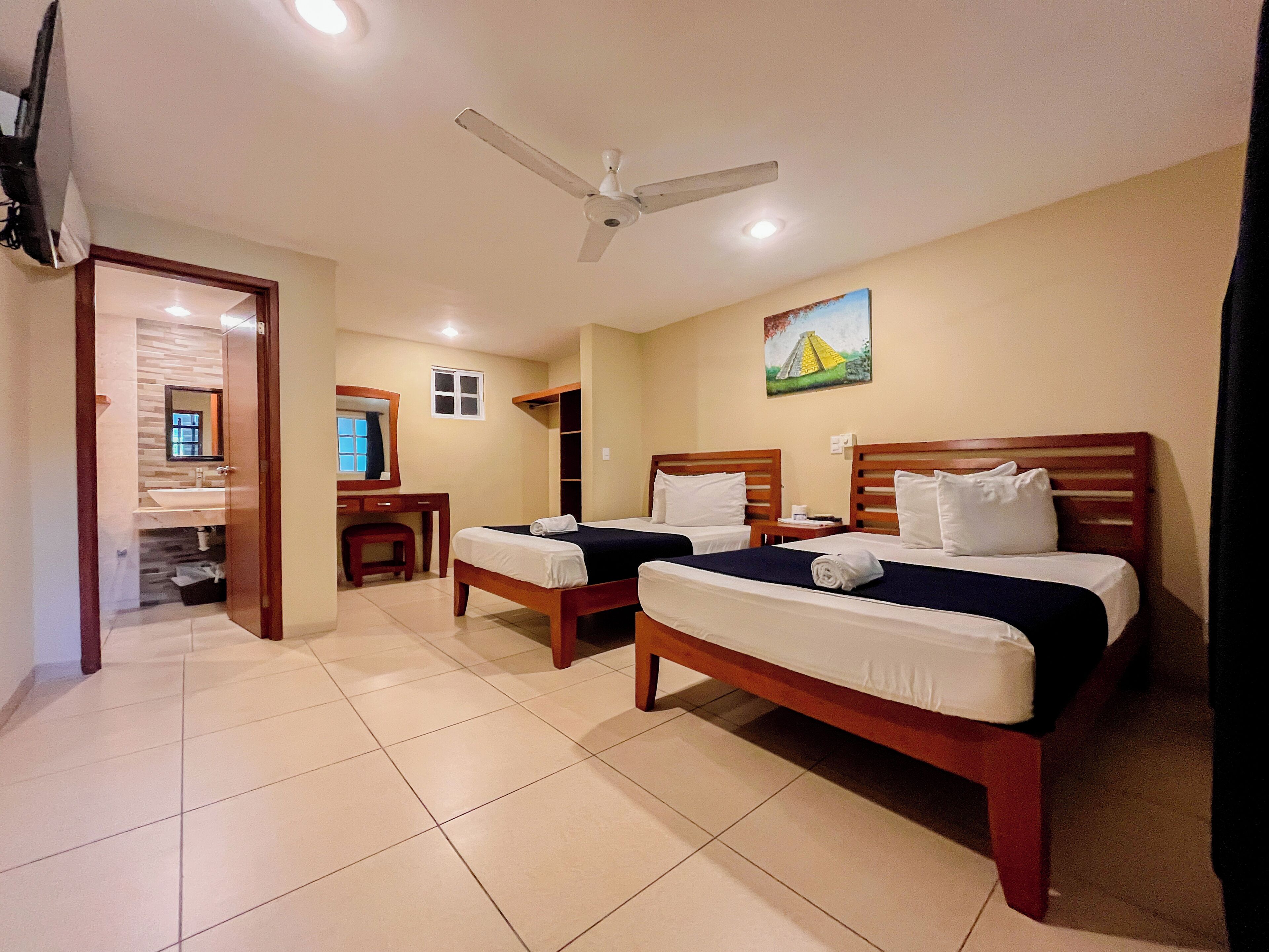 Habitación de hotel con dos camas, ventilador de techo, un cuadro y baño visible a través de una puerta abierta.
