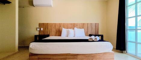 Una habitación de hotel con cabecera de madera, cama con ropa blanca, una mesita de noche pequeña, aire acondicionado de pared y una ventana con cortinas.