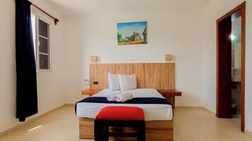 A hotel room with a bed, a wooden headboard, a painting on the wall, a window with curtains, and a door leading to a bathroom.