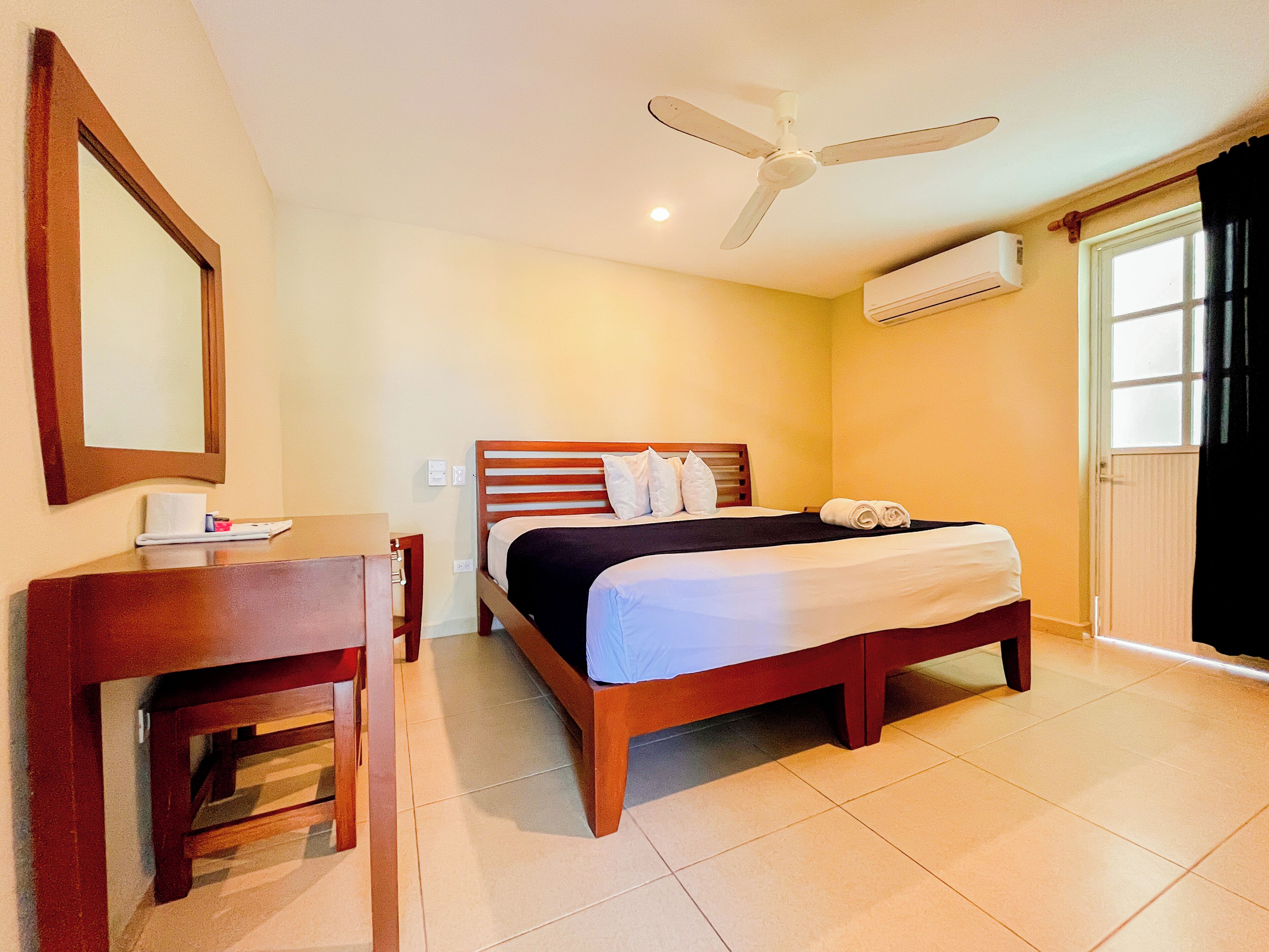 A hotel room with a bed, a desk, a chair, a ceiling fan, an air conditioning unit, and a window.