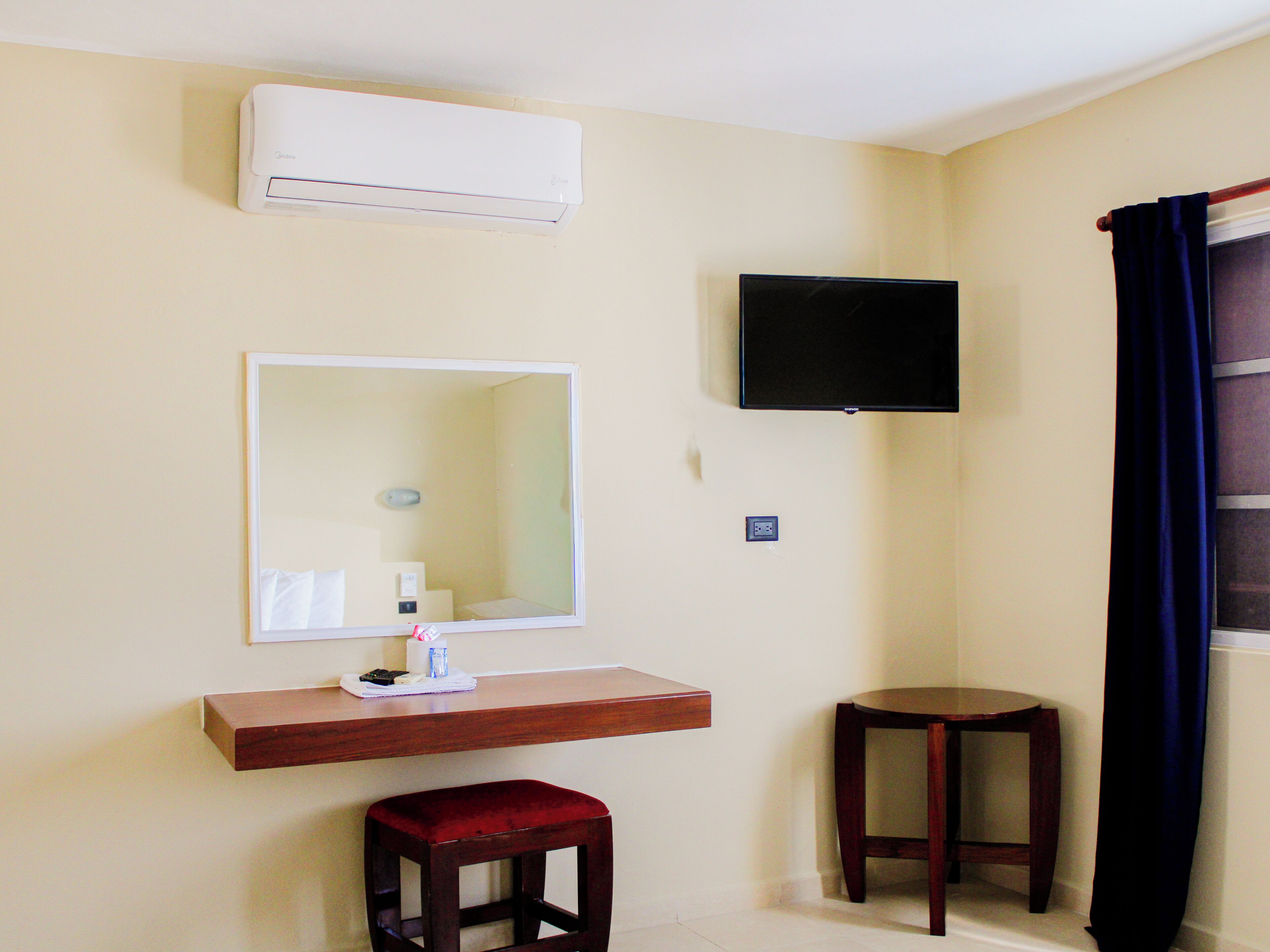 A room with a mounted TV, a mirror, a wooden shelf with toiletries, a red stool, and a small wooden table.