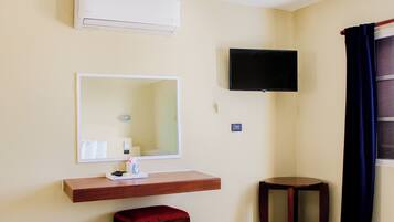 A room with a mounted TV, a mirror, a wooden shelf with toiletries, a red stool, and a small wooden table.