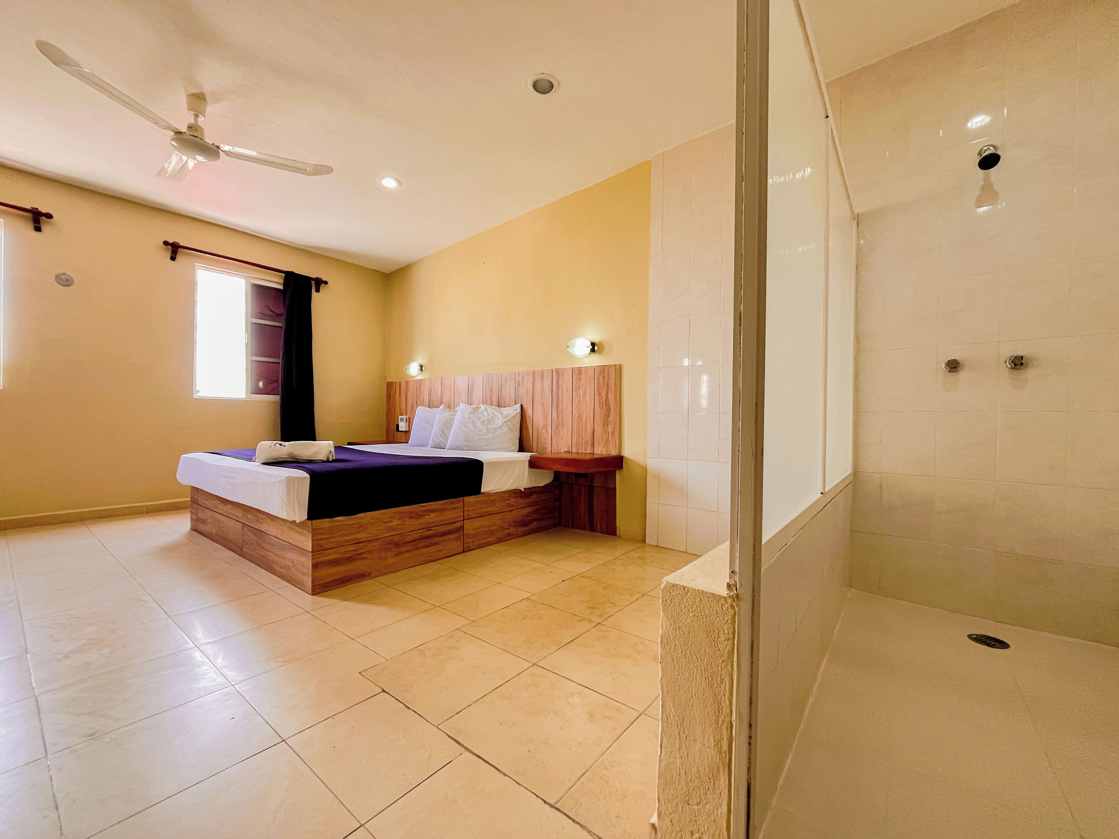 Un dormitorio con cama, ventilador de techo, ventana y zona de ducha.