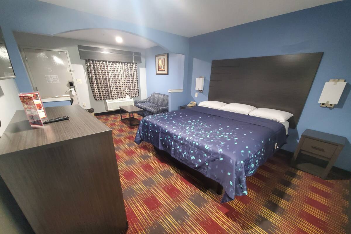 A hotel room with a bed, a desk, a chair, a small table, a wall-mounted heater, and a window with curtains.
