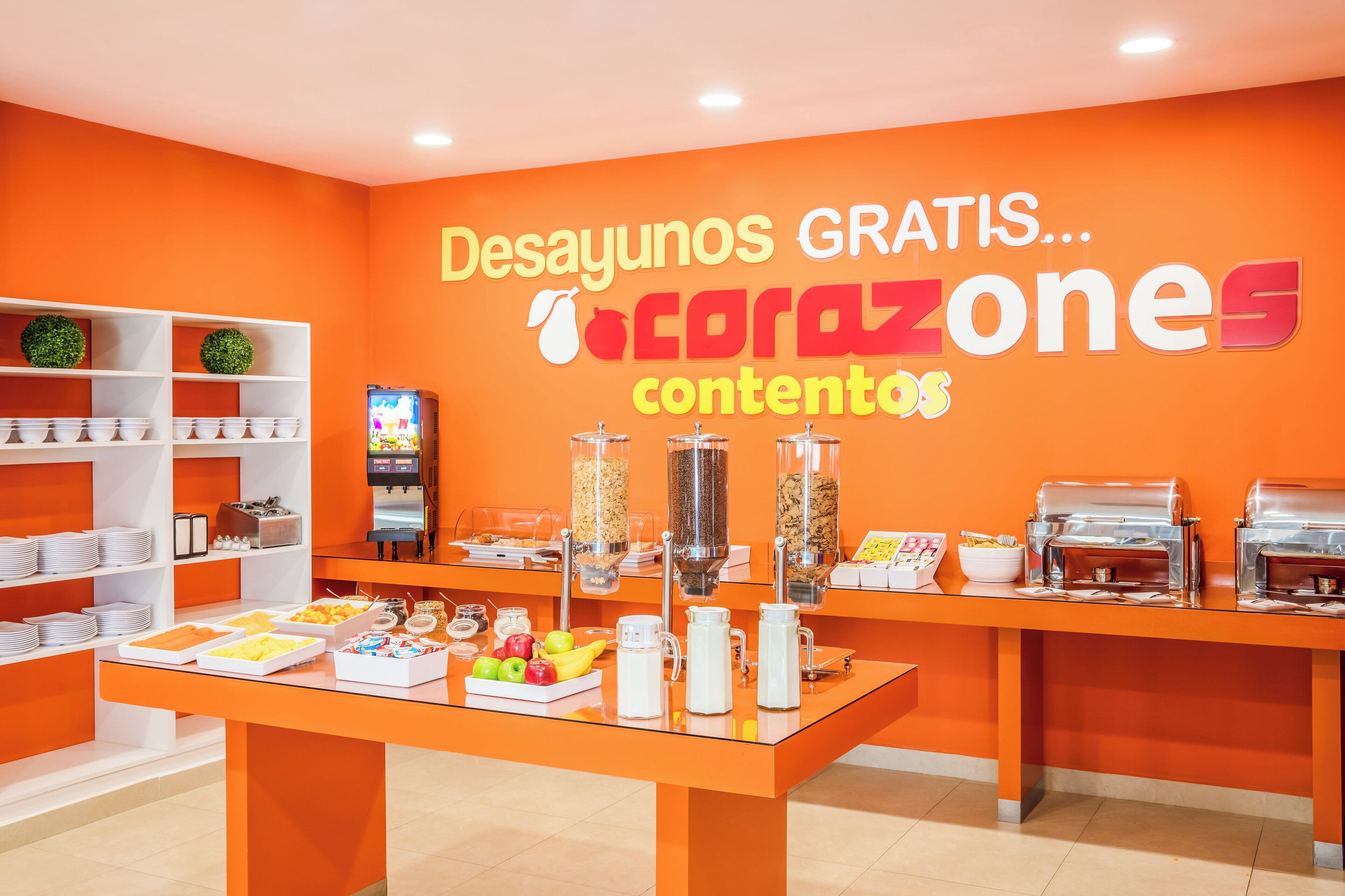 Espaço de buffet de café da manhã com paredes laranjas, prateleiras brancas e diversas opções de alimentos.