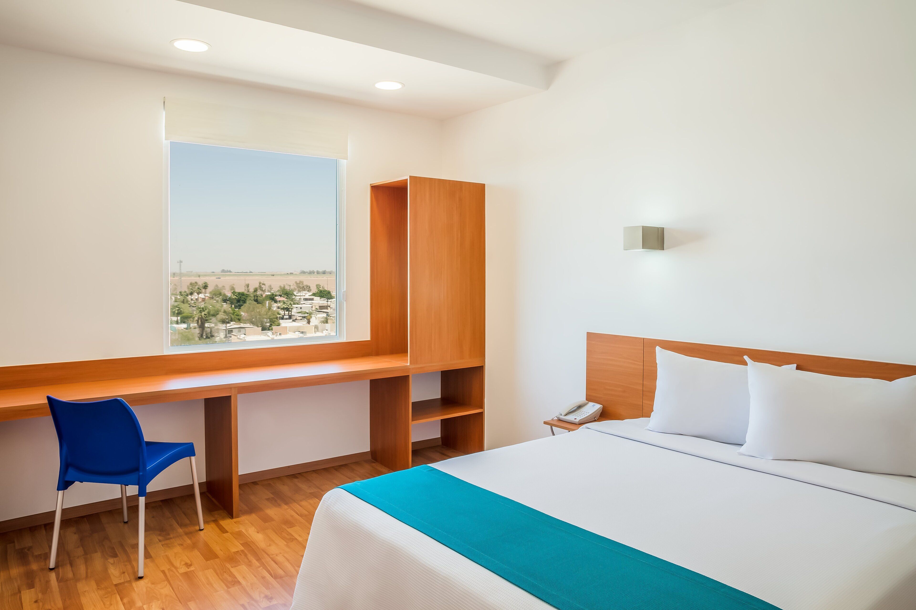 Habitación de hotel con cama, escritorio, silla, ventana con vista a la ciudad y teléfono en la mesita de noche.