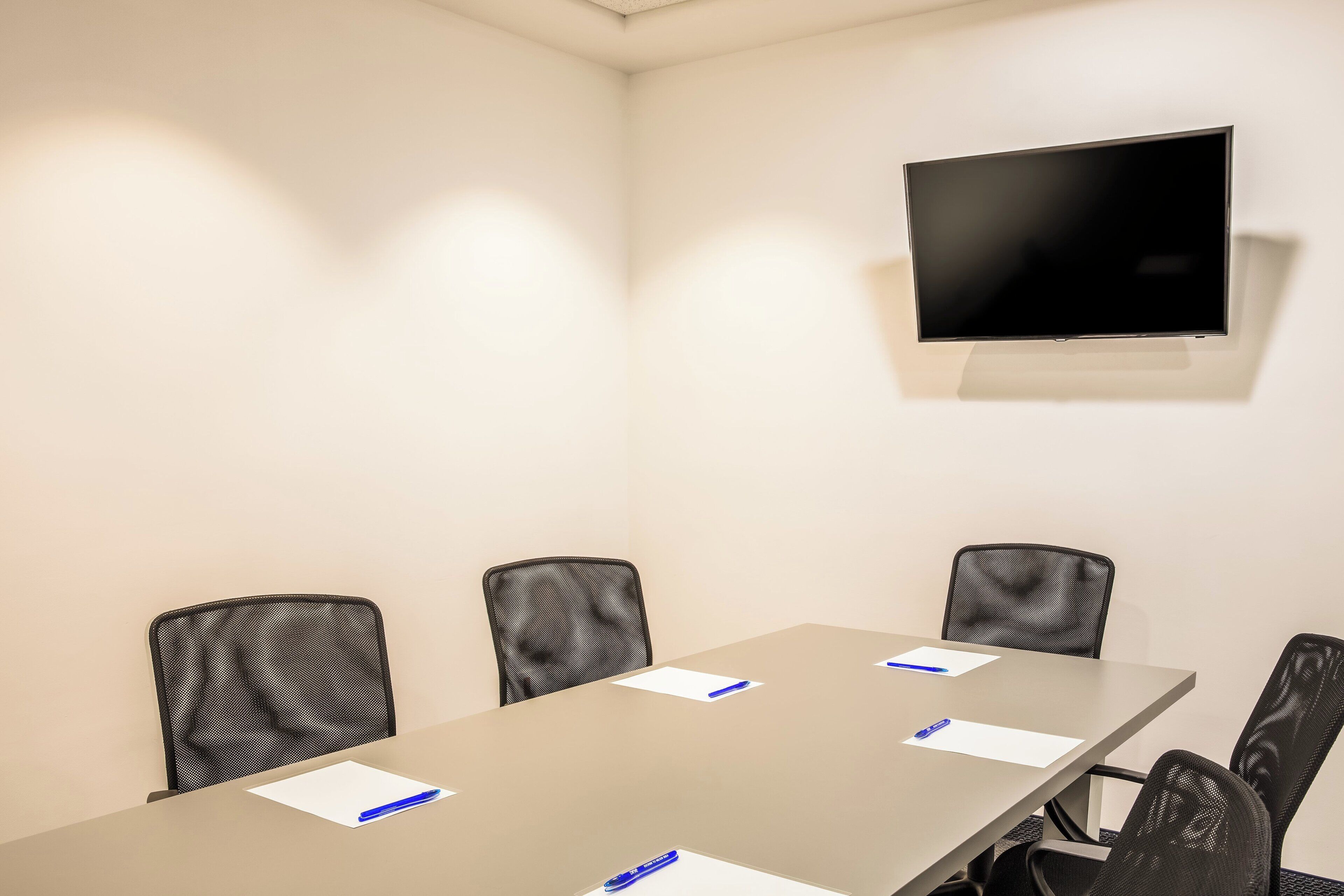 A conference room with a long table, four black chairs, a flat-screen TV mounted on the wall, and papers with pens on the table.