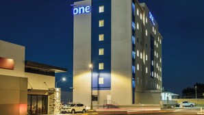 Un hôtel moderne, reconnaissable à son grand panneau lumineux représentant le chiffre « 1 », à sa façade en pierre et à son parking où sont stationnés des véhicules.