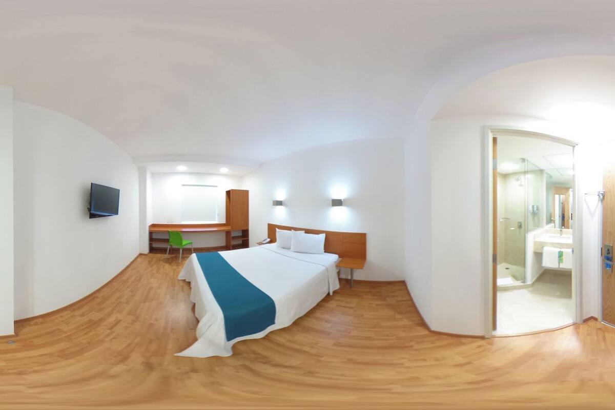 A bedroom with a bed, a desk, a chair, a television, and a bathroom visible through an open door.