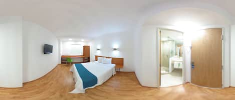 A modern hotel room with a wooden floor, a bed with a blue bedspread, a TV mounted on the wall, and a bathroom visible through an open door.