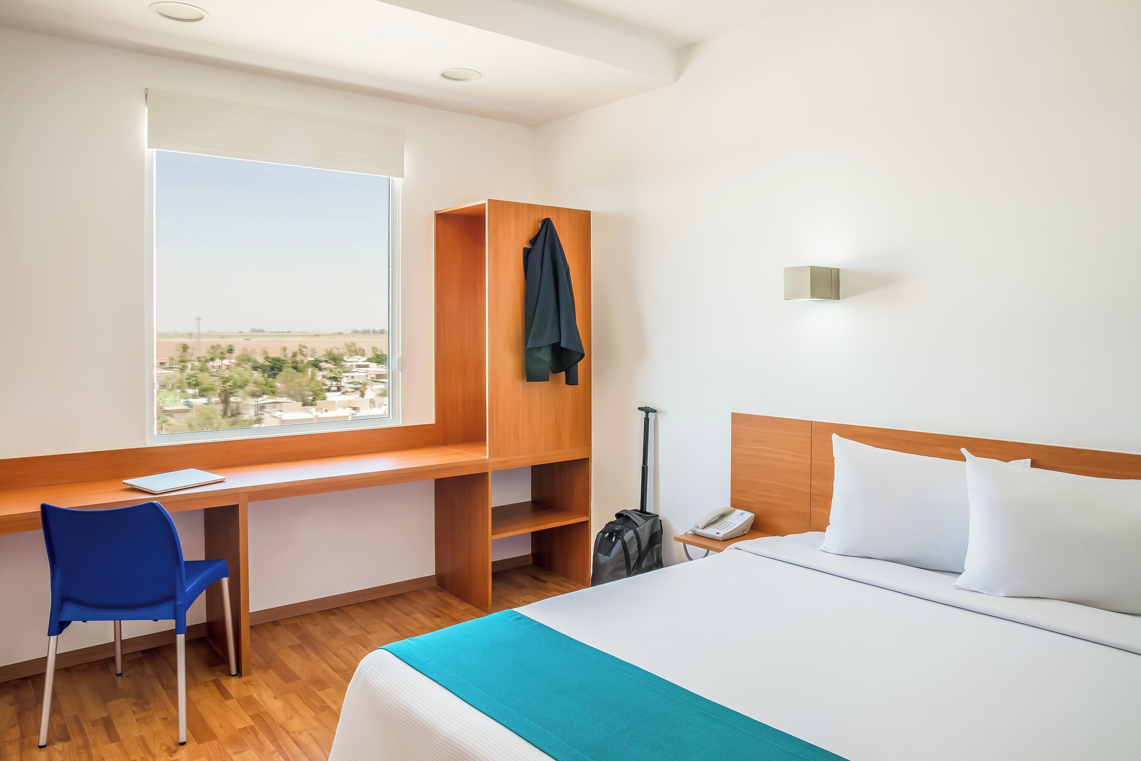 Una habitación de hotel con cama, escritorio, silla, maleta y vista a la ciudad.
