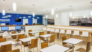 Une cafétéria moderne avec des tables et des chaises blanches, un mur bleu orné d’un téléviseur et un comptoir d’où l’on peut voir les voitures garées.