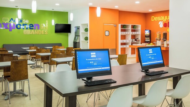 Un espace repas moderne avec des tables et des chaises, un ordinateur affichant un site web de réservation de voyages, et un mur aux couleurs vertes et oranges.