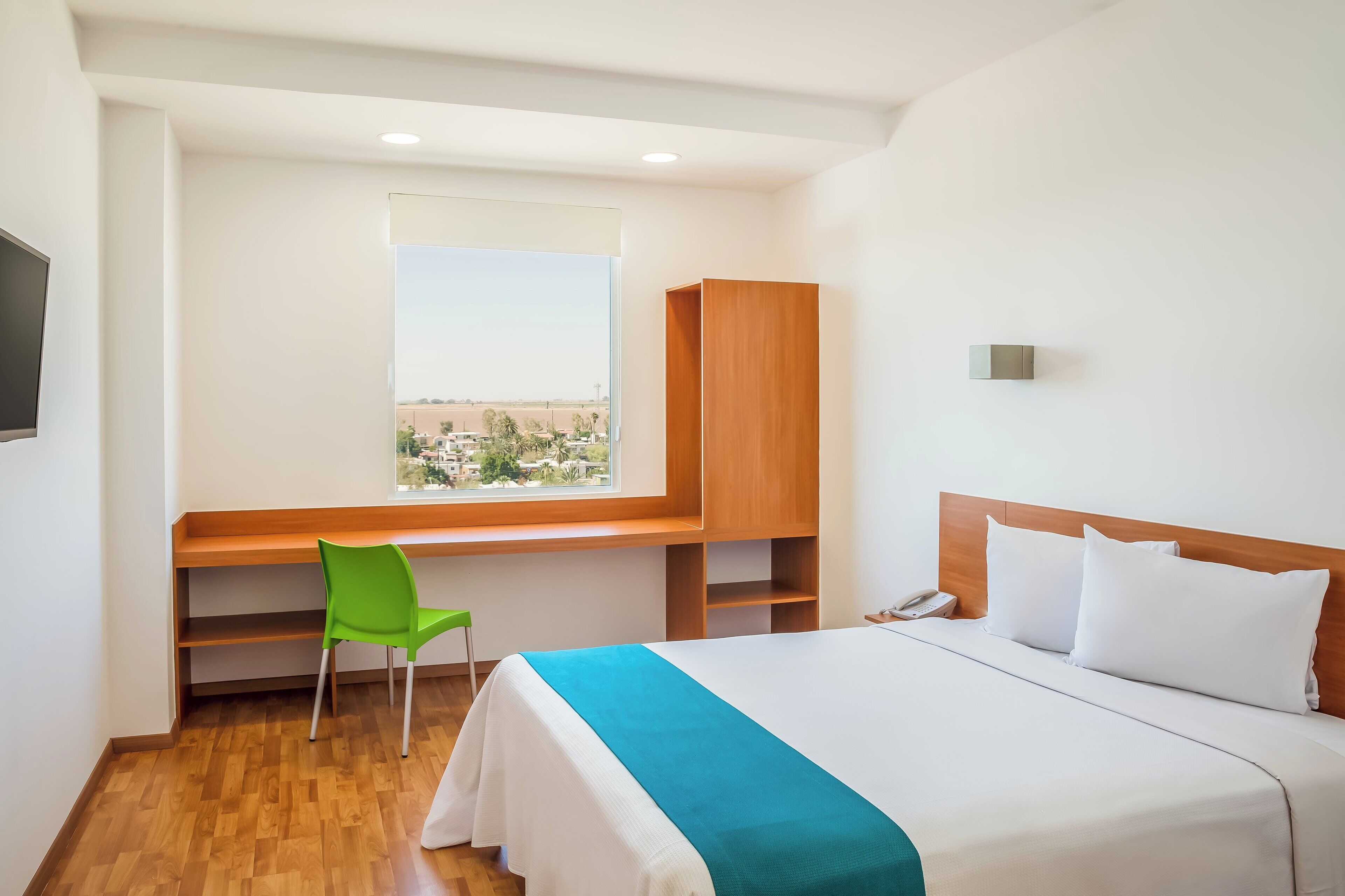 Habitación de hotel con cama, escritorio, silla, ventana con vistas y cabecero de madera.