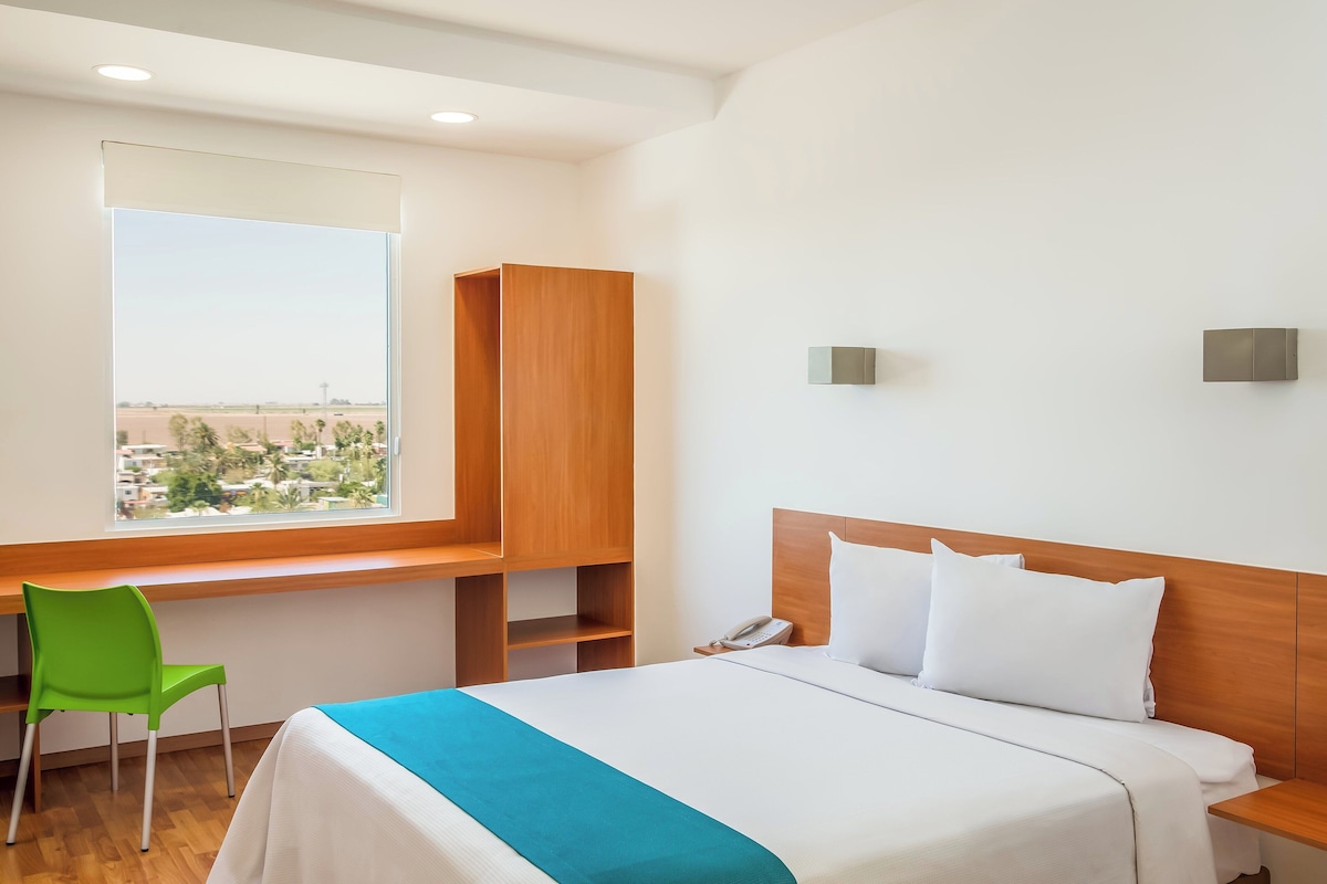 A hotel room with a bed, a desk, a chair, and a window with a view of the city.