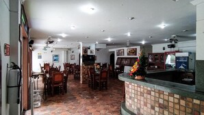 Un restaurante con suelo de baldosas, mesas y sillas de madera, una barra con planta decorativa y ventiladores de techo.